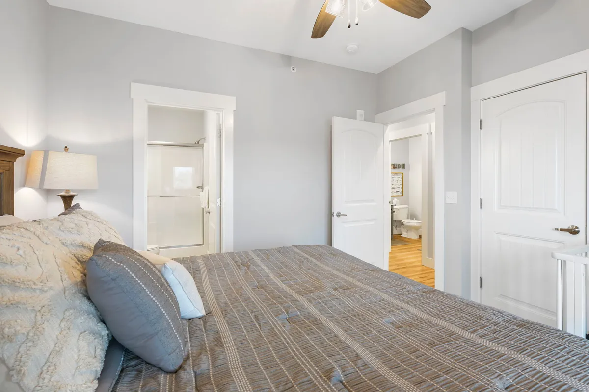 A neatly made bed with decorative pillows sits near open doors leading to an ensuite bathroom and hallway, creating a sense of spaciousness. Soft lighting and a bedside lamp enhance the cozy, restful vibe, while natural light streams into the room.