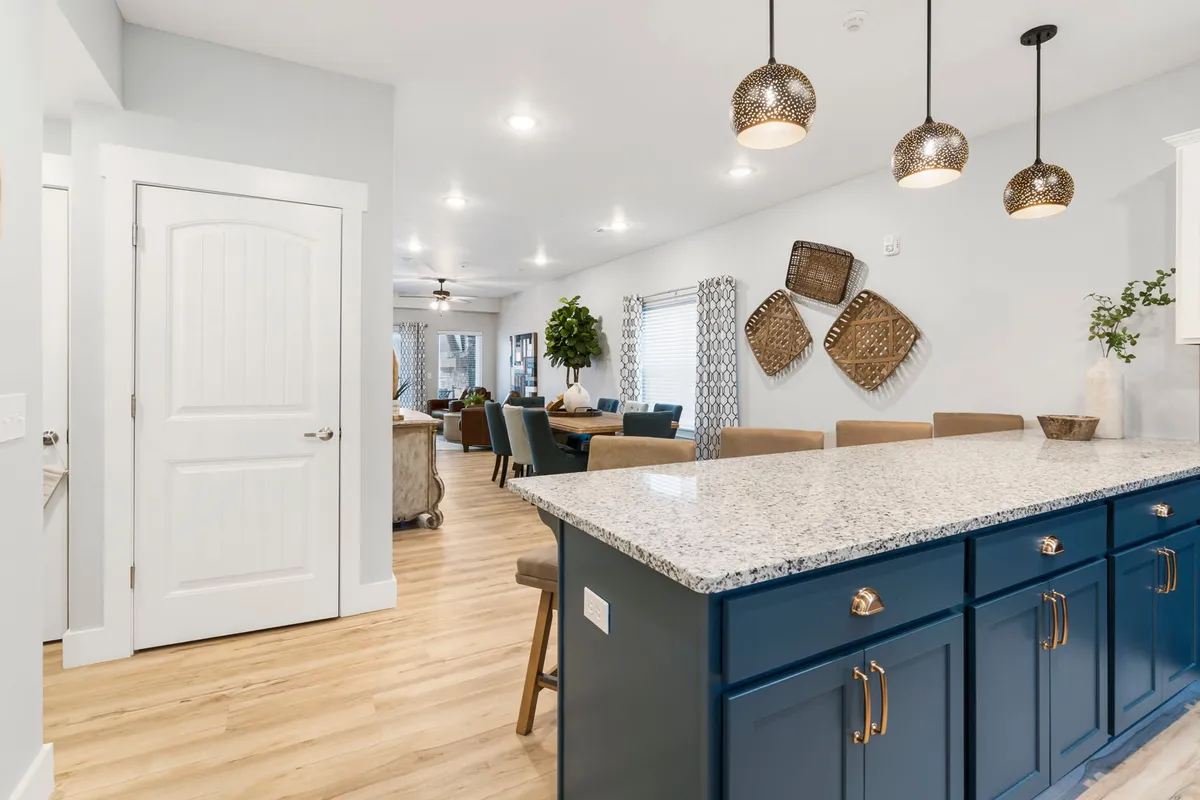 A sunlit, open kitchen featuring a large granite island, decorative pendant lights, and an open layout connecting seamlessly to the adjacent living and dining spaces. Stainless steel appliances and warm tones create an inviting atmosphere ideal for family cooking and gathering.