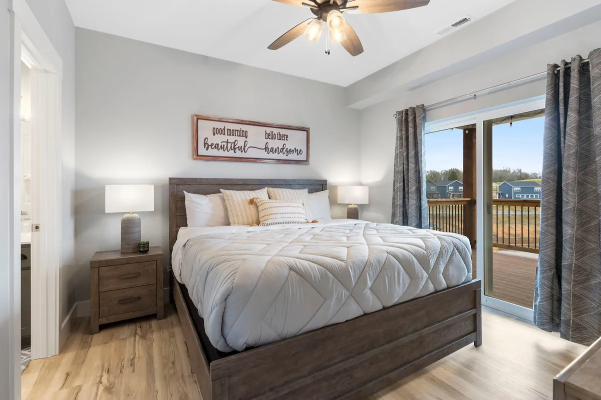 A king-sized bed with crisp white bedding is flanked by wooden nightstands and welcoming wall art. Large sliding doors open directly onto a balcony, flooding the room with natural light and offering seamless indoor-outdoor living. The space radiates tranquility and comfort.