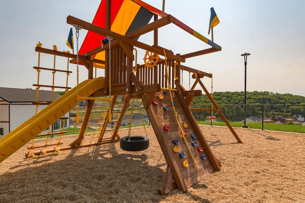 A vibrant playground featuring a colorful play structure with a climbing wall, slide, tire swing, and festive flags. Children are shown climbing, sliding, swinging, and playing together, creating an energetic and joyful scene.
