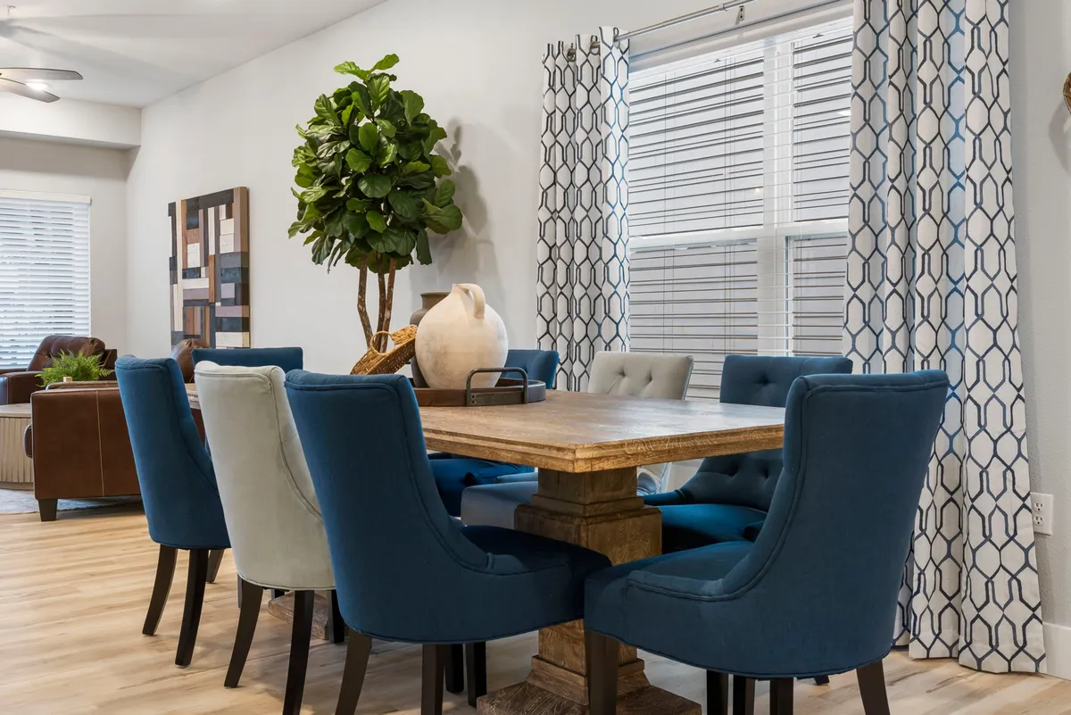 A welcoming dining area highlighted by a substantial table surrounded by blue chairs, a decorative plant centerpiece, and soft curtains that add warmth. The table is set for family meals and casual gatherings, emphasizing a sense of connection.