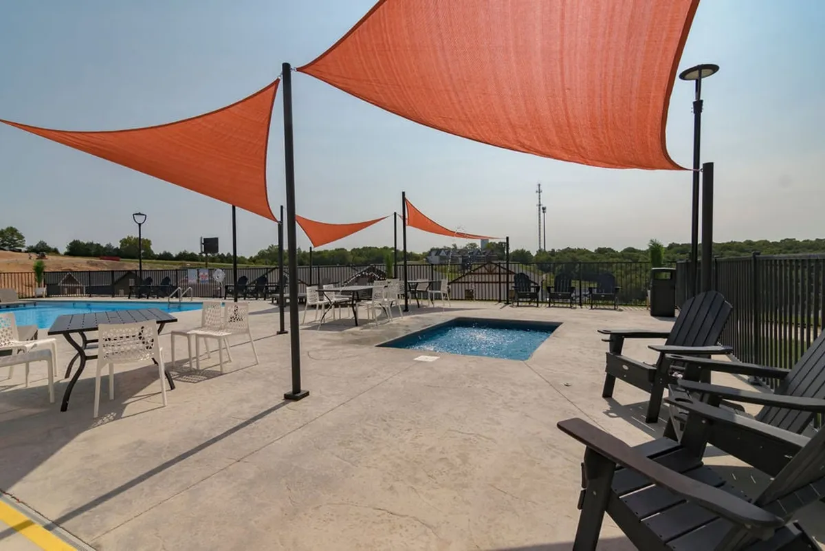 An inviting exterior pool scene with orange shade sails, Adirondack chairs, and space for swimming and social gatherings. The mood is leisurely and carefree, evoking group activities.