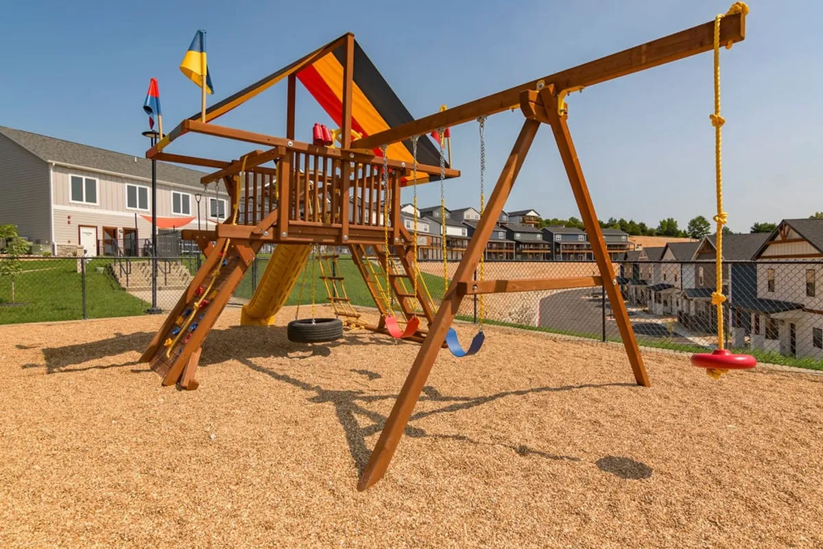 A joyful outdoor playground scene with children playing on a colorful structure and swings, surrounded by a sand surface and bright flags. The space encourages family interaction and outdoor activities.